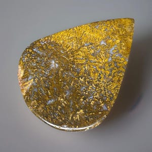May include: A teardrop-shaped gemstone with a golden-yellow and brown pattern resembling a forest. The stone has a polished surface and a translucent quality, with hints of white and gray throughout.