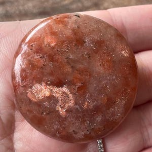 May include: A round, polished sunstone with a warm, reddish-orange hue and shimmering inclusions. The stone's surface is smooth and reflective, with a speckled appearance. The sunstone is a natural gemstone, perfect for jewelry or display.