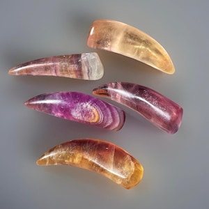 May include: Six polished fluorite crystal points in shades of purple, pink, and yellow-orange. The crystals have a smooth, curved shape, with visible banding and variations in color. The background is a solid gray.