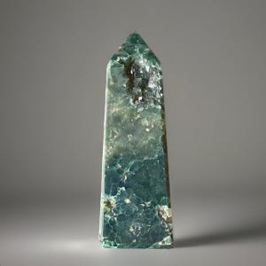 May include: A tall, rectangular crystal obelisk with a pointed top. The crystal is a mix of green and blue hues, with a textured surface and visible internal patterns. The obelisk stands upright against a neutral background.