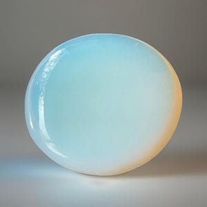 May include: A smooth, circular cabochon stone with a translucent appearance. The surface displays a gradient of soft light blue and milky white tones, transitioning to a warm, pale orange glow along the curved edge, reflecting light evenly.