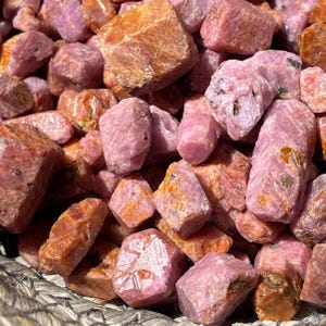 May include: A close-up shot of a collection of raw ruby gemstones. The stones vary in size and shape, with a mix of pink and orange hues. The rough texture of the stones is visible, suggesting they are in their natural, unpolished state. The gemstones are piled together.