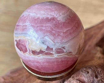 May include: A polished spherical rhodochrosite gemstone featuring concentric bands of pink, white, and cream colors. The stone is set within a circular gold-toned metal base, showcasing a smooth, glossy surface with natural mineral patterns.