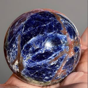 May include: A polished sodalite sphere, displaying a deep blue colour with white and reddish-brown veining. The gemstone ball is held in a hand, highlighting its smooth, spherical shape and natural patterns. A decorative item.