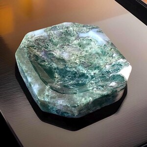 May include: A hexagonal-shaped, moss agate dish with a polished surface. The dish features a swirling pattern of green and white, with a translucent quality. The edges are faceted, and the dish sits on a dark surface.