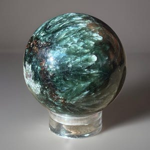 May include: A spherical gemstone, polished to a high sheen, displaying a captivating pattern of dark green and brown. The orb rests on a clear, cylindrical stand. The stone's surface reflects light, accentuating its unique textures and natural beauty.