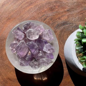 May include: A bowl filled with amethyst crystals, the purple stones are various sizes and shapes, the bowl is clear glass and sits on a wooden surface.