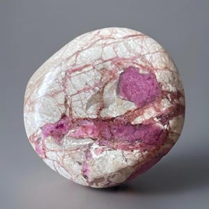 May include: A polished, oval-shaped stone with a cream-colored base and striking pink and magenta veining and inclusions. The stone has a smooth, rounded surface and a natural, organic appearance. The pink hues are concentrated in irregular patterns throughout the stone.