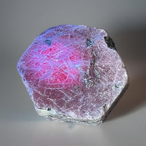 May include: A rough-cut gemstone with a hexagonal shape. The stone has a deep pink core with a network of white lines and a purplish-gray exterior. Small black inclusions are visible on the surface. The gem is set against a neutral background.