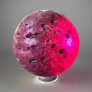 May include: A decorative sphere with a textured surface, illuminated with a gradient of pink and purple hues. The sphere has several dark, crater-like indentations and sits on a clear, circular base.