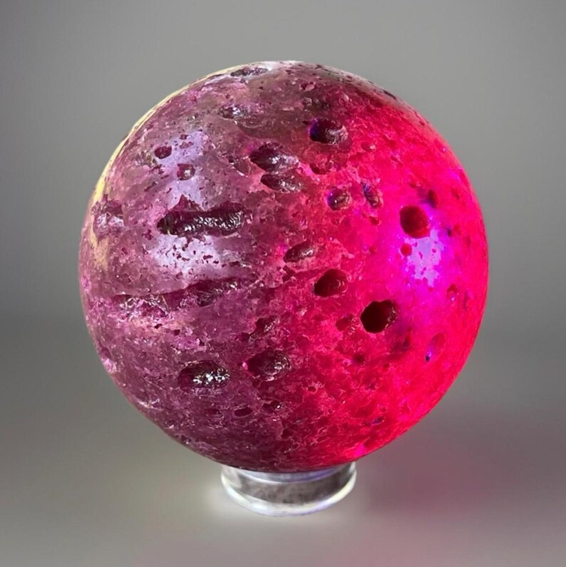 May include: A decorative sphere with a textured surface, illuminated with a gradient of pink and purple hues. The sphere has several dark, crater-like indentations and sits on a clear, circular base.
