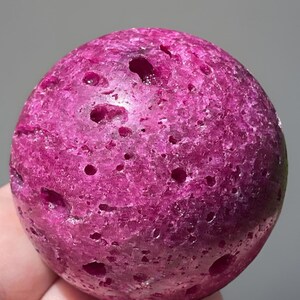 May include: A close-up of a spherical, magenta-colored stone with numerous small holes. The stone has a rough, textured surface and appears to be a natural mineral or gemstone. The lighting highlights the stone's color and unique features.