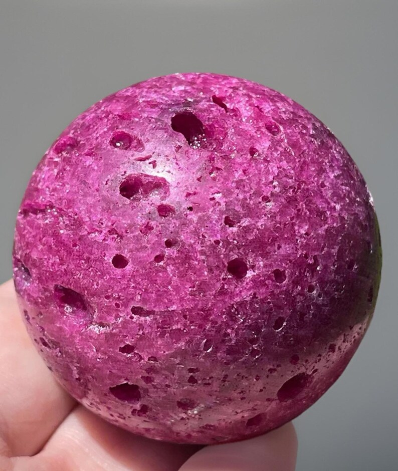 May include: A close-up of a spherical, magenta-colored stone with numerous small holes. The stone has a rough, textured surface and appears to be a natural mineral or gemstone. The lighting highlights the stone's color and unique features.
