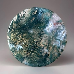 May include: A round, polished moss agate stone with a swirling pattern of green, white, and tan colors. The stone has a smooth, glossy surface and a natural, organic appearance. The agate is a decorative item.