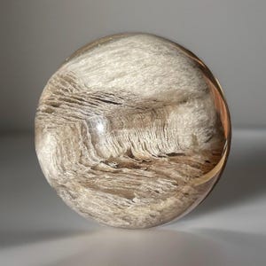 May include: A clear crystal ball with a layered, textured interior. The sphere has a light brown and white color scheme, with a polished, reflective surface. The interior layers create a unique, natural pattern.