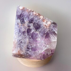 May include: A large, raw amethyst crystal with a polished, angled edge. The crystal displays a range of purple hues, from deep violet to pale lavender, with areas of white and reddish-brown. It rests on a small, round wooden base.