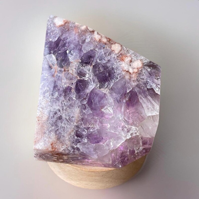 May include: A large, raw amethyst crystal with a polished, angled edge. The crystal displays a range of purple hues, from deep violet to pale lavender, with areas of white and reddish-brown. It rests on a small, round wooden base.