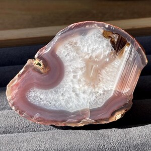 May include: A polished agate stone slice with a natural, organic shape. The stone features concentric bands of colour, including white, lavender, and reddish-brown hues. The centre has a crystalline appearance, and the edges are smooth and translucent.