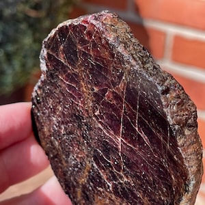 May include: A raw, uncut gemstone with a deep burgundy color and a network of white veins. The stone has a rough, textured surface and appears to be naturally formed. The gemstone is held in a hand.