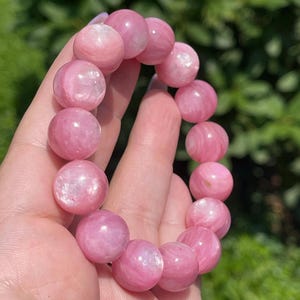 May include: A pink gemstone bracelet featuring large, round beads. The beads have a soft, translucent appearance with subtle variations in color. The bracelet is held against a blurred green background, highlighting the pink tones of the stones.