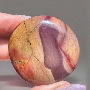 May include: A round, polished stone with a mix of colors. The stone features a large, curved section of deep purple, contrasted by tan, red, and yellow hues. Fine cracks and veins add texture to the surface. The stone is held between fingers.