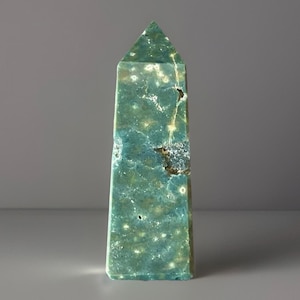May include: A tall, green obelisk-shaped crystal with a pointed top. The stone has a mottled pattern of teal, light green, and cream colors. The crystal is standing upright against a gray background.