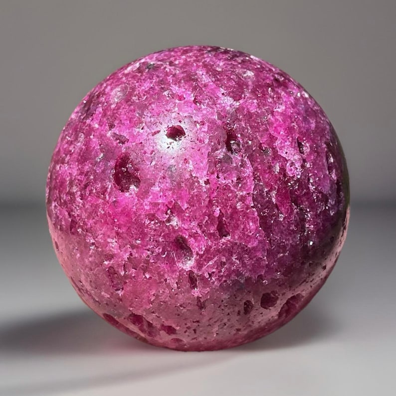 May include: A spherical, polished gemstone in shades of deep pink and magenta. The surface has a textured, crystalline appearance with small, natural indentations. The gemstone is a solid, rounded shape, and the color is consistent throughout.