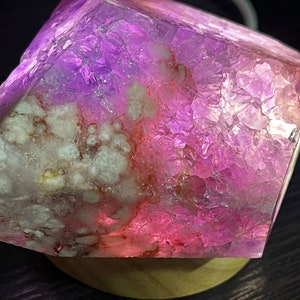 May include: A faceted, cube-shaped crystal with a gradient of pink and purple hues. The crystal sits on a round, wooden base. The interior of the crystal has white and red inclusions. The light refracts through the crystal, creating a glowing effect.