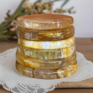 May include: A stack of seven translucent, golden-yellow gemstone bangles. The bangles have a rounded, flat shape and vary slightly in colour, with some having white and brown inclusions. The stack is on a white lace doily.
