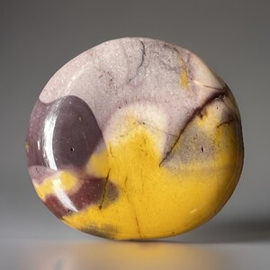 May include: A polished, rounded stone with a striking color palette. The stone features a vibrant yellow base, overlaid with patches of deep purple and pale lavender. Dark brown veins add further detail to the natural patterns.