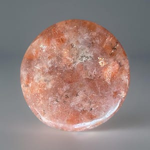 May include: A round, polished sunstone gemstone with a mix of orange, red, and white hues. The stone has a sparkling, reflective quality, with small inclusions and a translucent appearance. The gemstone is set against a neutral background.