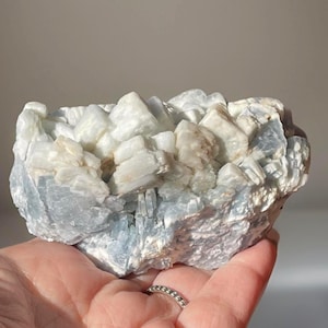 May include: A close-up of a cluster of light blue and white crystal formations. The crystals have a rough, textured appearance, with varying sizes and shapes. The overall colour palette is soft and natural, with pale blue and creamy white hues.