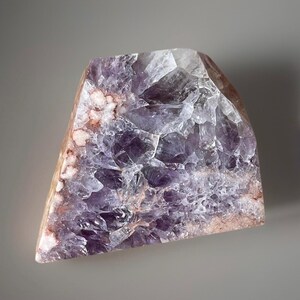 May include: A large, raw amethyst crystal with a triangular shape. The crystal features a mix of deep purple, clear, and light brown hues. The surface has a rough, textured appearance, showcasing the natural formation of the stone.
