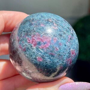 May include: A polished sphere of blue-green stone with vibrant pink inclusions. The gemstone ball has a smooth, reflective surface, showcasing the natural patterns and colors within the stone. The sphere is held in a hand, highlighting its size and shape.