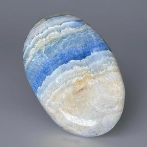 May include: A polished, oval-shaped stone with a layered appearance. The stone features bands of white, light blue, and deep blue hues, creating a natural, ocean-like aesthetic. The stone is smooth and appears to be a decorative item.