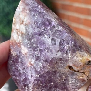 May include: A polished amethyst crystal with a pointed top. The crystal displays shades of purple, white, and brown. The surface has a crystalline structure with visible facets. The stone is held in a hand.