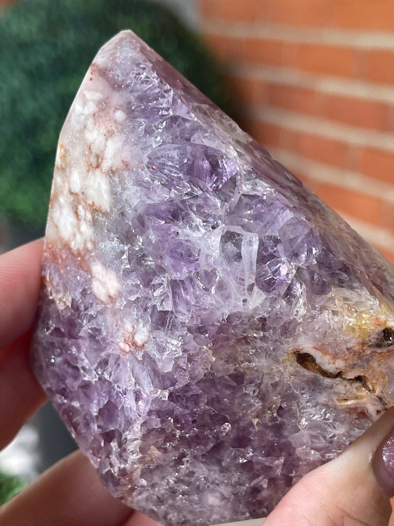 May include: A polished amethyst crystal with a pointed top. The crystal displays shades of purple, white, and brown. The surface has a crystalline structure with visible facets. The stone is held in a hand.