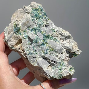 May include: A rough, gray rock formation with patches of vibrant green and blue mineral deposits. The stone has a textured surface with visible crystalline structures, showcasing natural geological beauty. The rock is held in a hand.