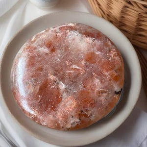 May include: A round, polished sunstone crystal with orange and white hues, displayed on a beige plate. The sunstone has a sparkling, translucent appearance, with visible inclusions. The crystal is a natural, decorative item.