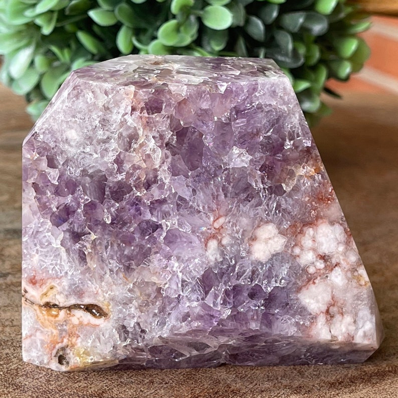May include: A large, raw amethyst crystal geode with a faceted top. The crystal features a deep purple hue with white and orange inclusions. The rough-cut edges and natural texture are visible.