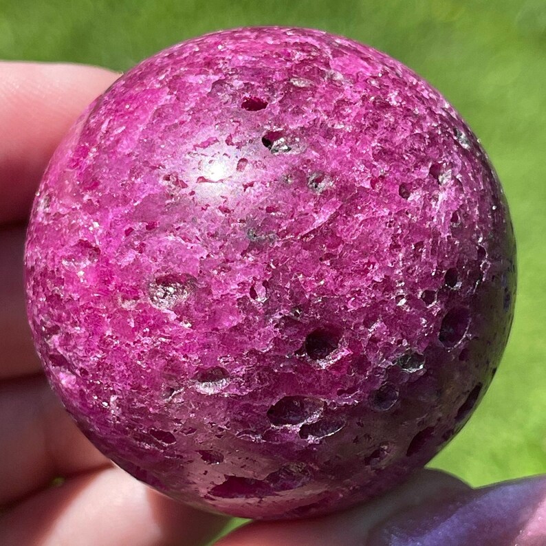 May include: A close-up of a polished, spherical gemstone in a vibrant magenta hue. The surface has a textured appearance with small, natural holes. The sphere is held against a blurred green background, highlighting its color and form.