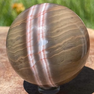 May include: A polished spherical stone featuring horizontal bands of earthy brown and tan tones, intersected by a prominent vertical stripe of translucent white and soft pink mineral layers that wrap around the smooth, rounded surface.