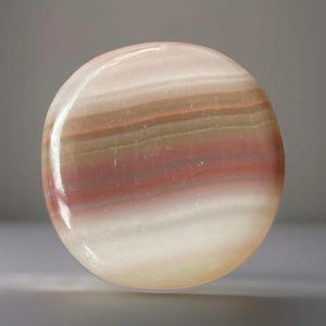 May include: A polished, round stone with a layered appearance. The stone features bands of white, light pink, brown, and red hues. The surface is smooth and reflective, with a slight sheen. The overall shape is a perfect circle.