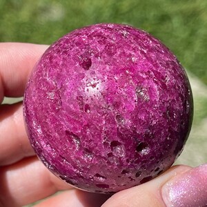 May include: A vibrant, polished sphere of deep magenta-pink stone. The surface has a textured appearance with natural variations in color and small, irregular pits. The sphere is held in a hand, with a blurred green background.