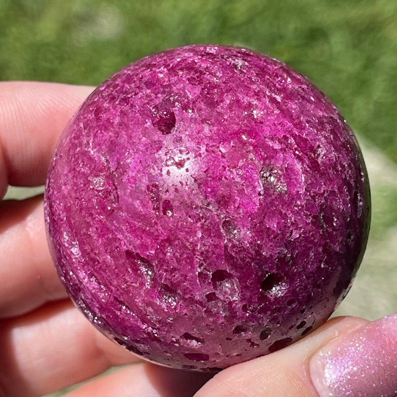 May include: A vibrant, polished sphere of deep magenta-pink stone. The surface has a textured appearance with natural variations in color and small, irregular pits. The sphere is held in a hand, with a blurred green background.