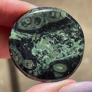May include: A round, polished stone with a dark green and black pattern. The stone has a swirling design with lighter green and silver flecks throughout. The stone is held between two fingers.