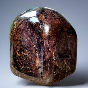 May include: A polished, rounded gemstone with a deep burgundy color and a glossy surface. The stone has a complex internal structure with visible veins and inclusions, creating a unique pattern. The overall shape is smooth and organic, with a slight translucence.