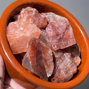 May include: A collection of raw, rough-cut red quartz crystals fills a small, round, orange-brown ceramic bowl. The crystals vary in size and have a translucent appearance with reddish-orange and clear inclusions. The bowl is smooth and has a slightly glossy finish.