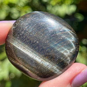 May include: A polished, oval-shaped gemstone with a dark, metallic appearance. The stone has a mix of dark gray and brown tones with shimmering, reflective inclusions. The surface is smooth and rounded, with a subtle sheen.