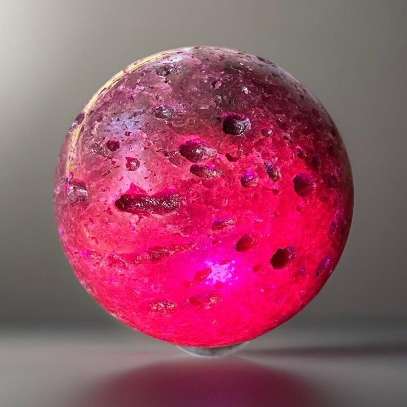 May include: A glowing, spherical object with a textured surface. The sphere transitions from a deep magenta at the base to a purplish-brown at the top, with several small holes. The object appears to be illuminated from within, creating a vibrant, otherworldly effect.